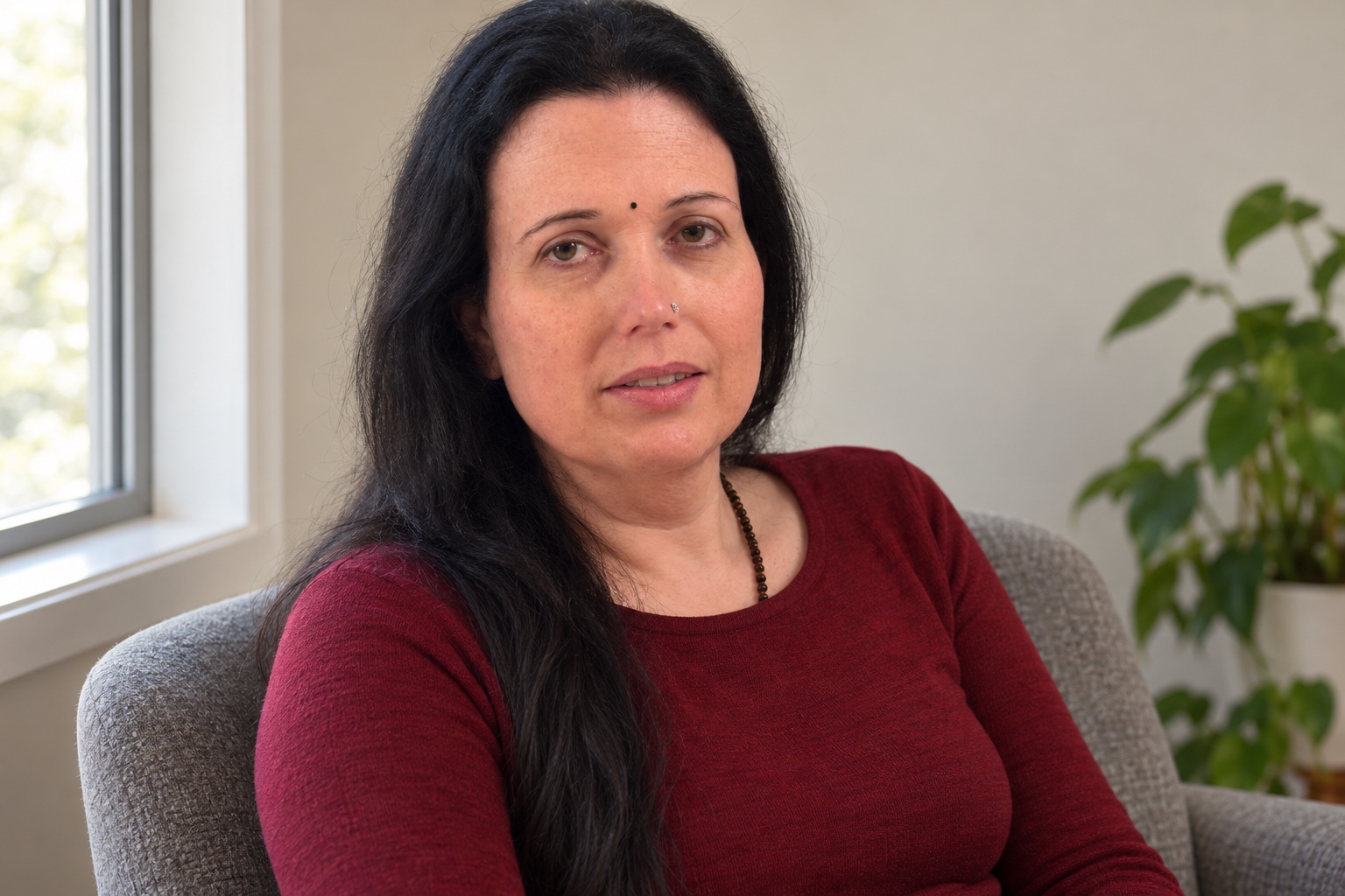 Prem Devi seated beside a window in a grey chair, wearing a dark red top, small black bindi and left-side nose pin